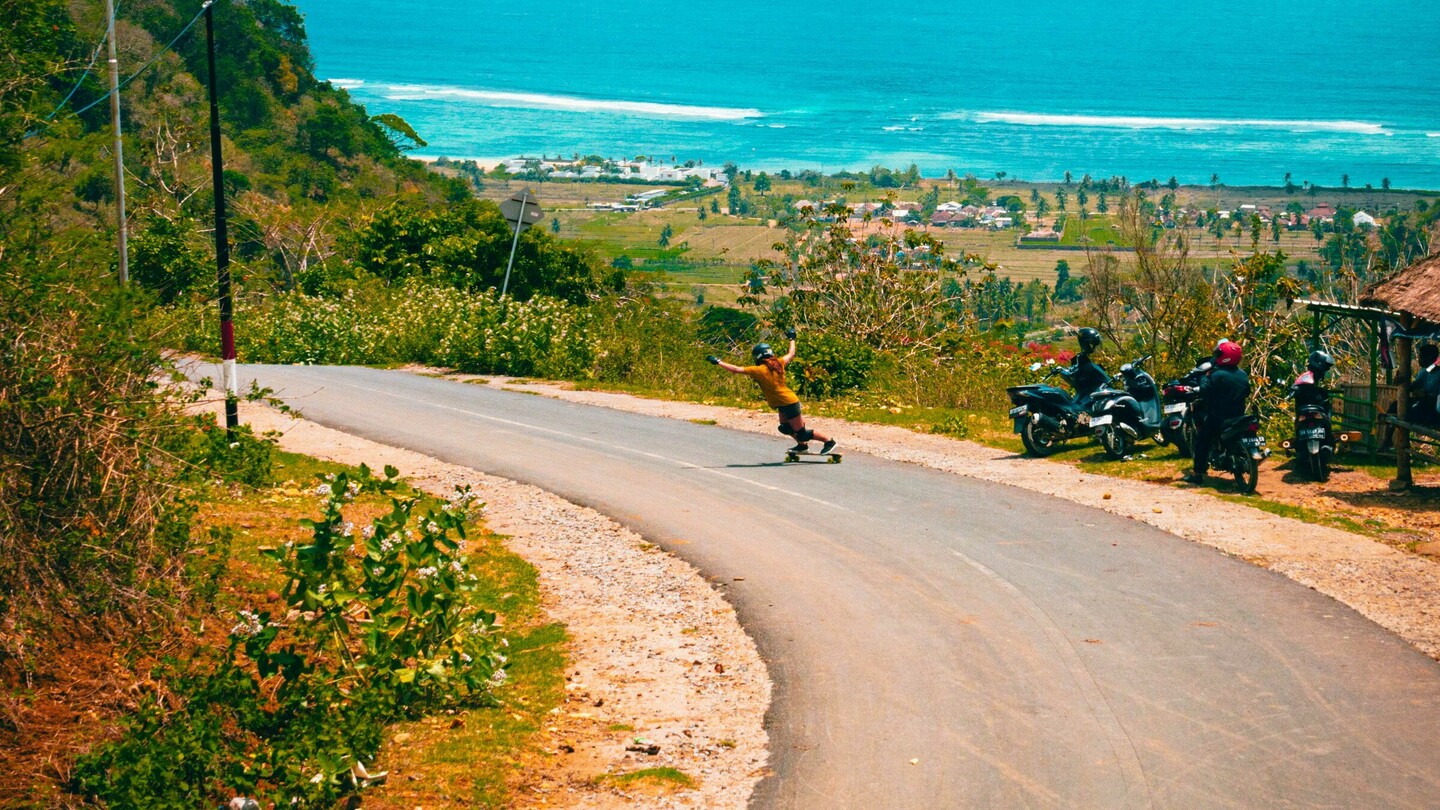 A downhill skateboarder speeds down a winding road that leads to a stunning coastal view with turquoise blue sea. The road winds through green scenery, flanked by vegetation and a few motorcycles on the roadside. In the background, the ocean stretches out under a bright blue sky with white clouds, adding a summery and adventurous vibe to the scene.