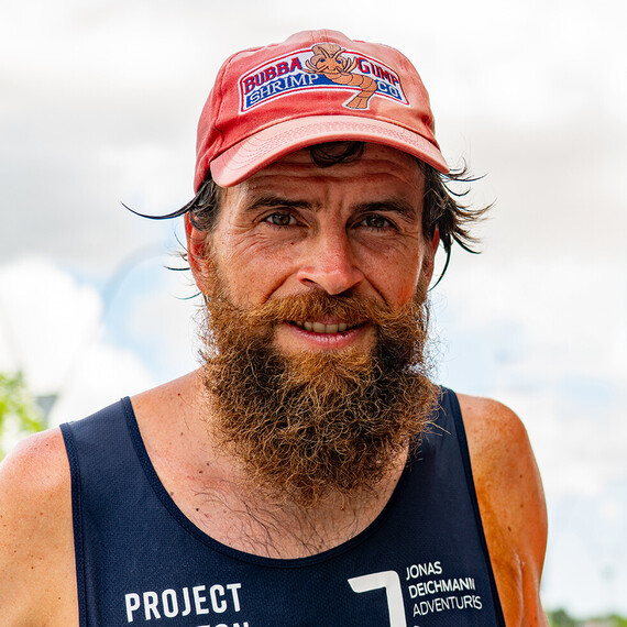 Portrait of Jonas Deichmann, a man with a thick beard and red cap featuring the Bubba Gump Shrimp Co. logo. He wears a dark tank top.